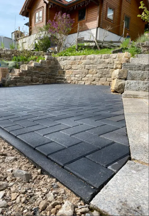 Frisch verlegte Pflasterfläche aus dunklem Betonstein vor einer Natursteinmauer